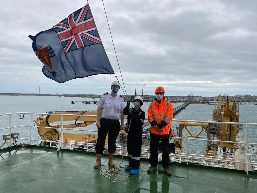 El Capitán Ralph Stevens, la marinera Paula Muñoz y el Director de Operaciones del BAS, Simon Garrod, izan la bandera azul que lleva el escudo modificado del escudo de armas del Territorio Antártico Británico. Foto: Capitán Will Whatley