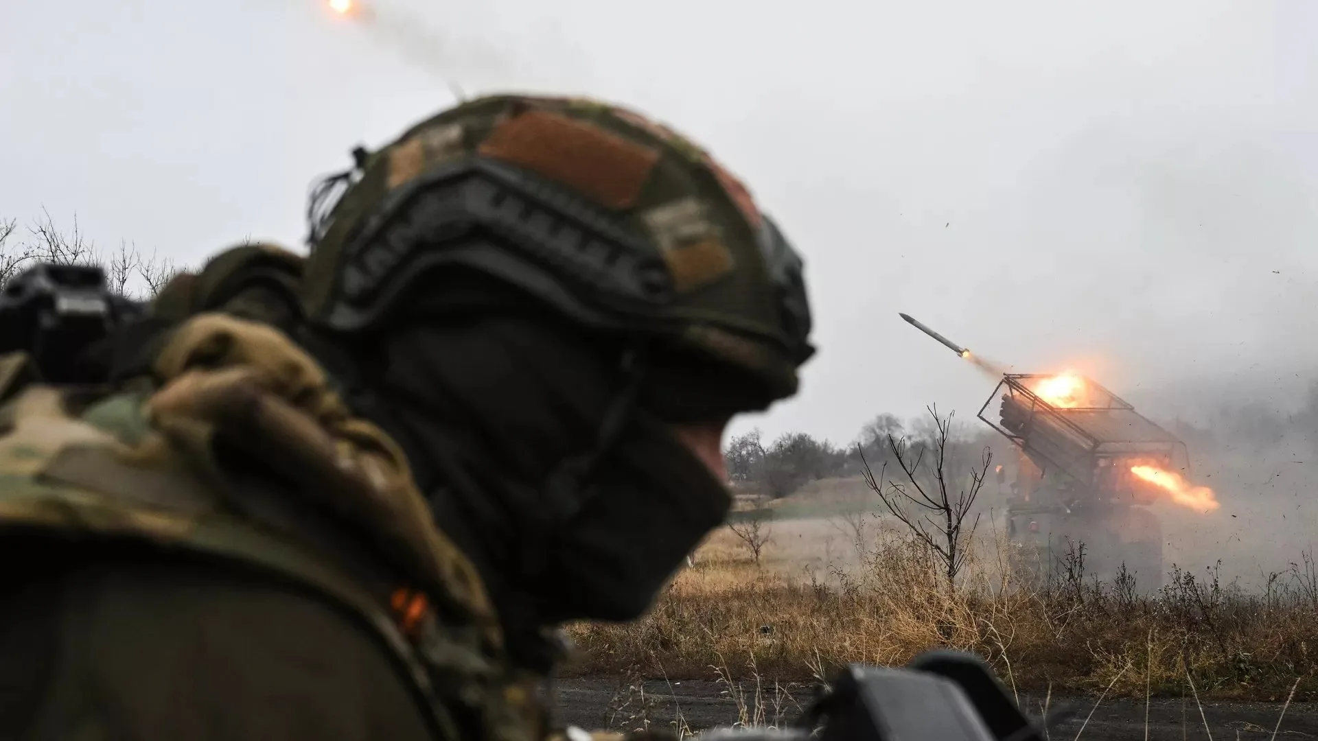 Un sistema de lanzacohetes múltiple (MLRS) efectúa un disparo en zona de operaciones mientras un militar ruso permanece en primer plano durante la secuencia de lanzamiento/ Créditos: Sputnik / Stanislav Krasilnikov.