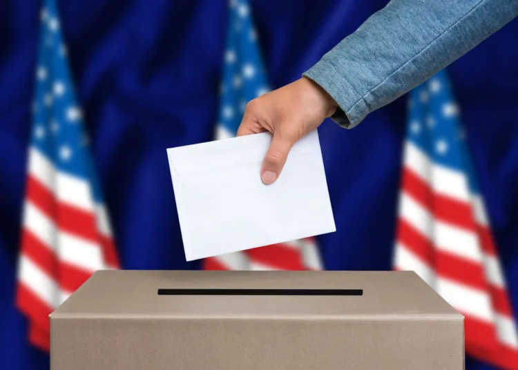 Presidential election in United States of America. The hand of woman putting her vote in the ballot box. American flags on background.
