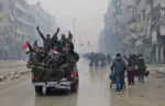 Syrian pro-regime fighters, gesture as they drive past residents fleeing violence in the restive Bustan al-Qasr neighbourhood, in Aleppo's Fardos neighbourhood on December 13, 2016, after regime troops retook the area from rebel fighters. 
Syrian rebels withdrew from six more neighbourhoods in their one-time bastion of east Aleppo in the face of advancing government troops, the Syrian Observatory for Human Rights said. / AFP PHOTO / STRINGER