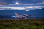 Un caza J-10 despegando en pista con postquemador encendido (llama azul), fondo montañoso con turbinas eólicas. Pertenece a una brigada de aviación de la Fuerza Aérea del Comando Sur del EPL. Créditos: China Military Bugle.