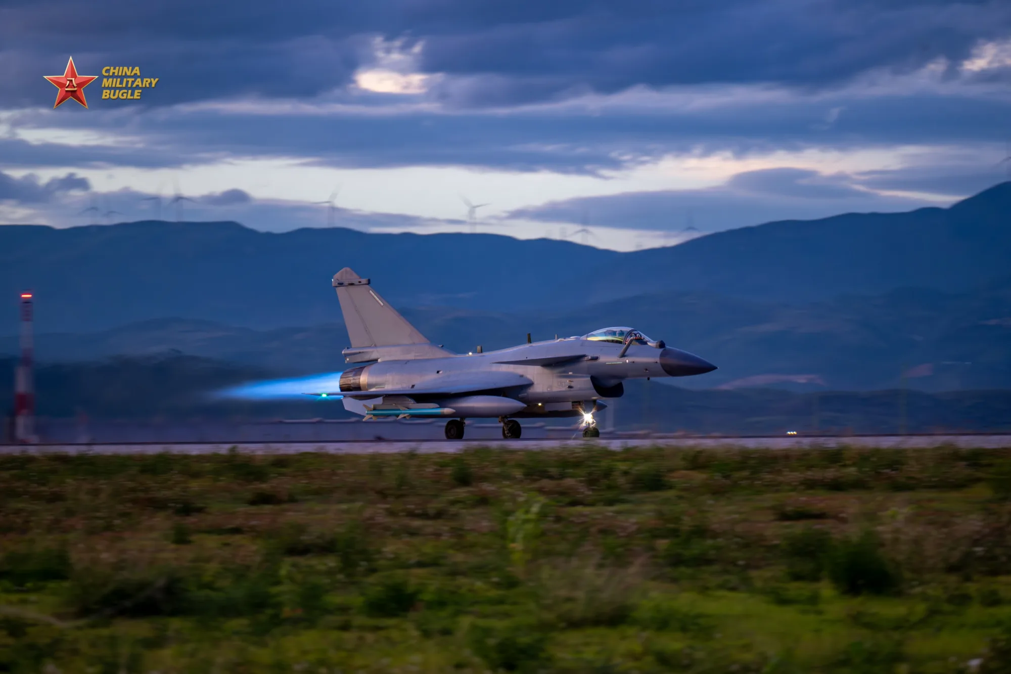Un caza J-10 despegando en pista con postquemador encendido (llama azul), fondo montañoso con turbinas eólicas. Pertenece a una brigada de aviación de la Fuerza Aérea del Comando Sur del EPL. Créditos: China Military Bugle.