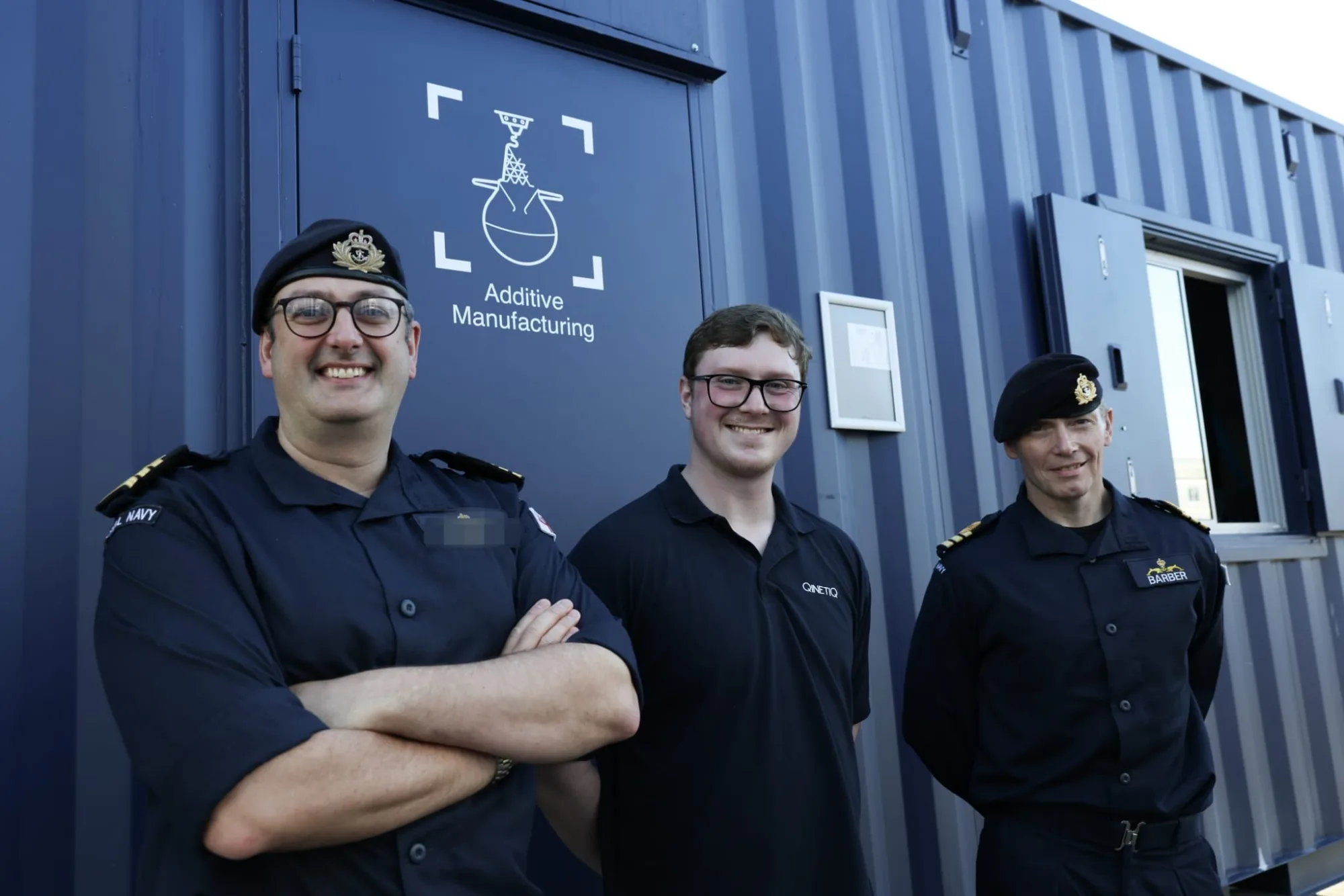 Personal naval junto a expertos de QinetiQ frente a contenedores con el logo de Additive Manufacturing. Créditos: Royal Navy in Scotland.